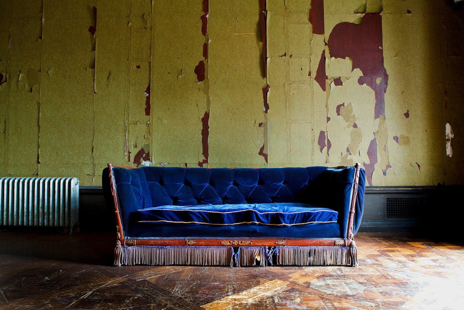 Blue velvet sofa against peeling wallpaper on a worn parquet floor at Alder Manor
