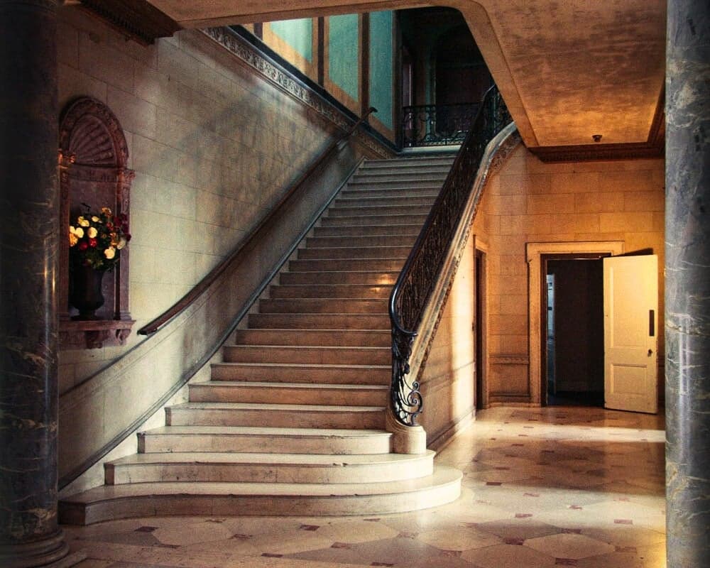 Marble staircase with a wrought-iron railing in a stone hall at Alder Manor, flanked by marble columns