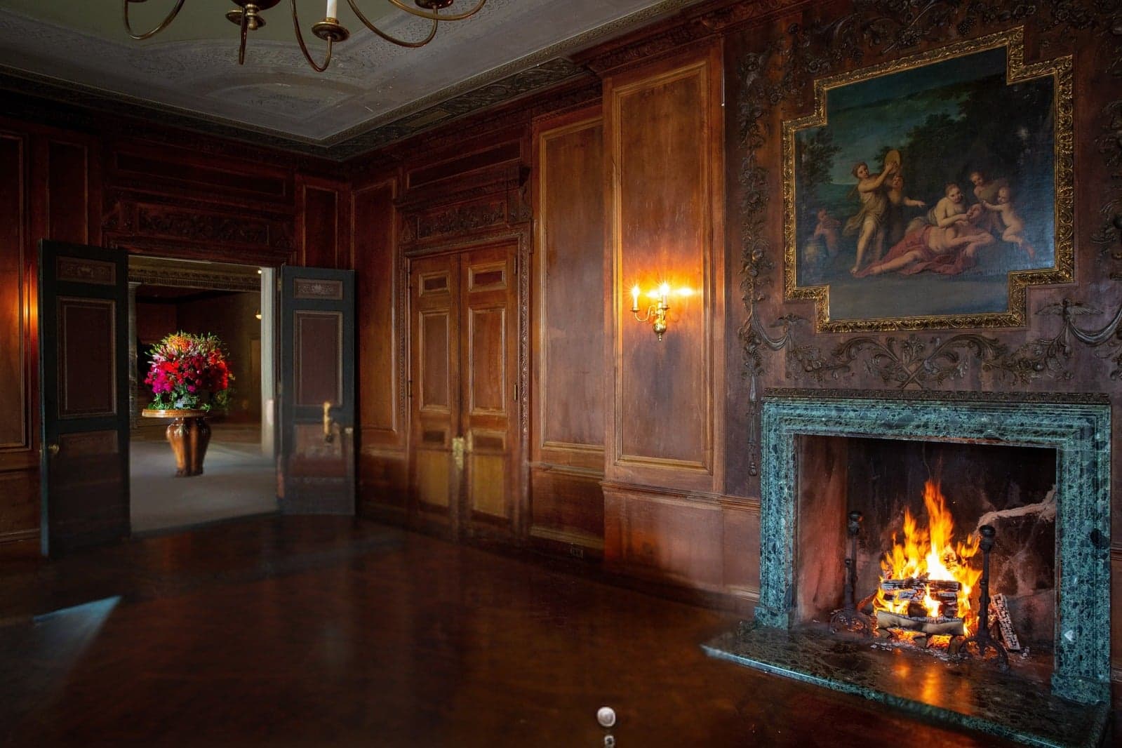 Wood-paneled room at Alder Manor with a fire in a green marble fireplace and an oil painting above it