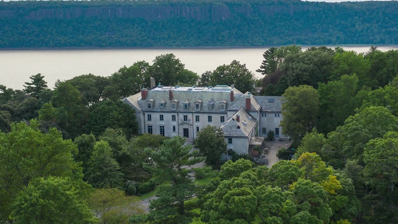 Alder Manor from the air, a limestone mansion among green trees above the Hudson River, with the cliffs of the Palisades across the water