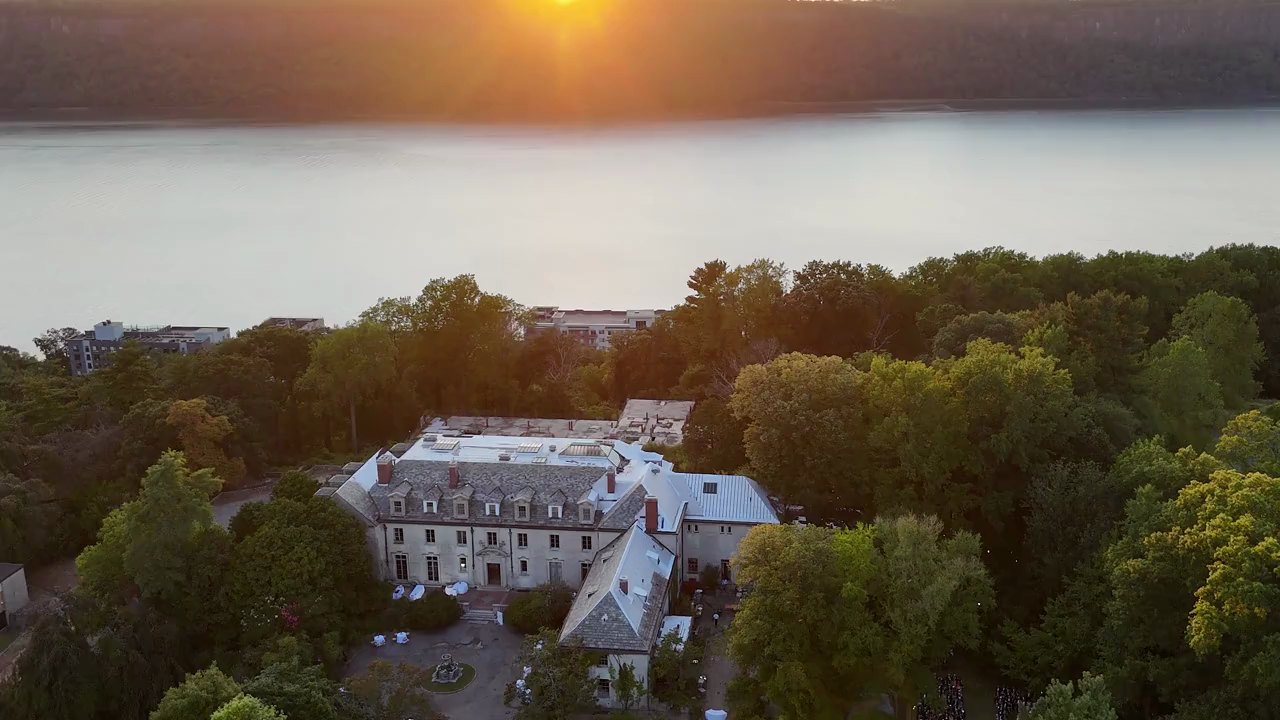 Alder Manor among the trees above the Hudson River at sunset, with the sun going down over the Palisades across the water